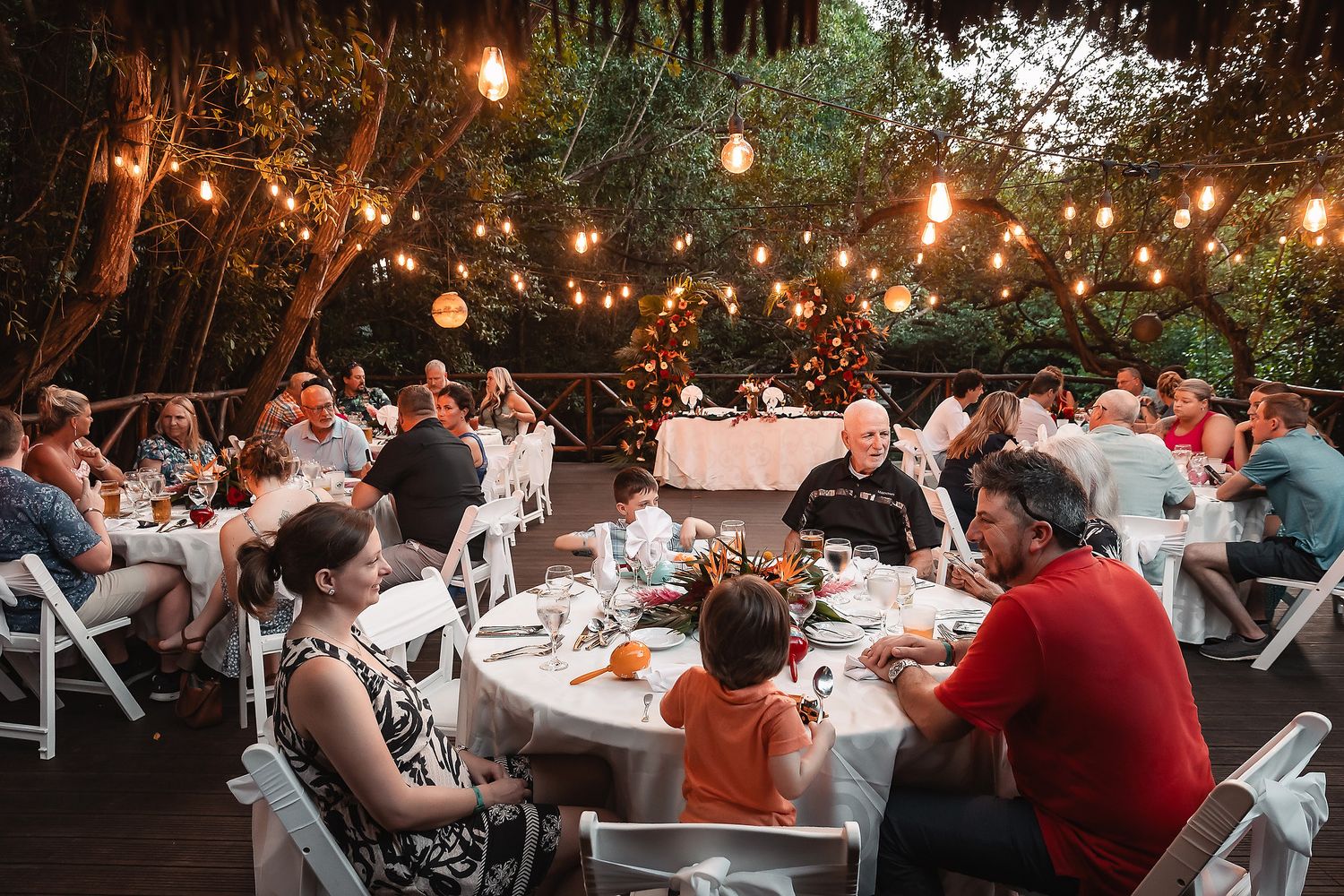 Outdoor wedding reception at dusk with string lights and guests dining at round tables.