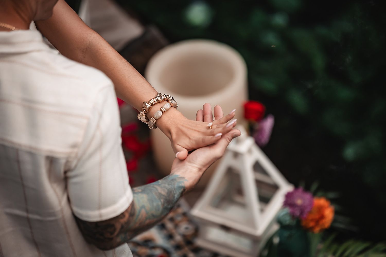 Close-up of hands with jewelry and tattoos against a blurred outdoor background with white lantern.