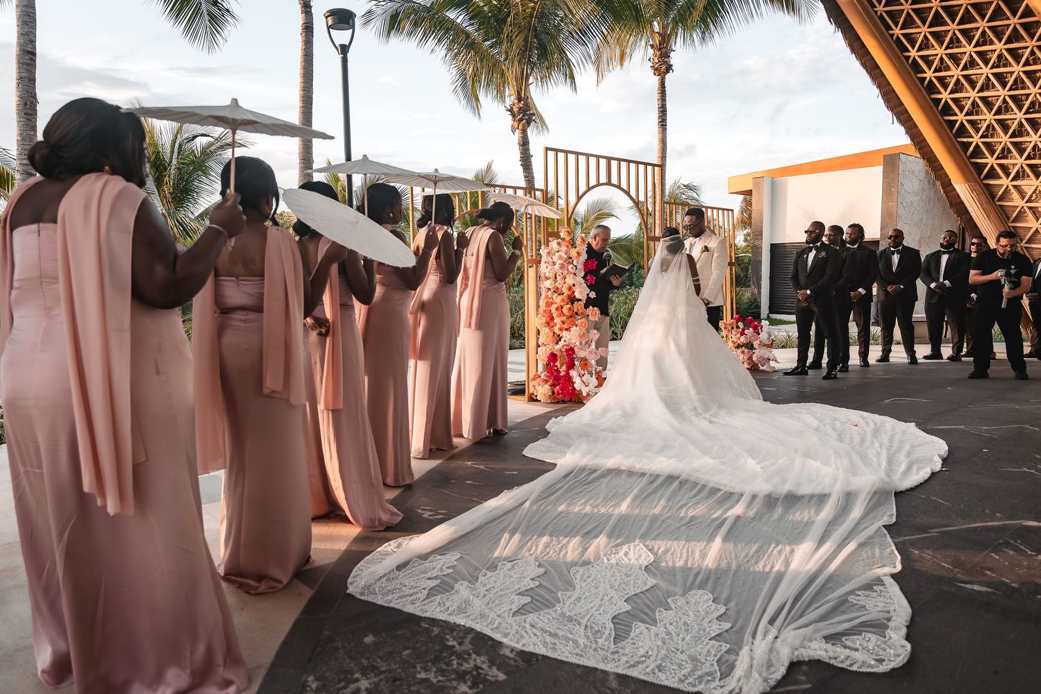 Bridesmaids in pink dresses stand beside a bride with a flowing cathedral veil at a tropical outdoor wedding ceremony.