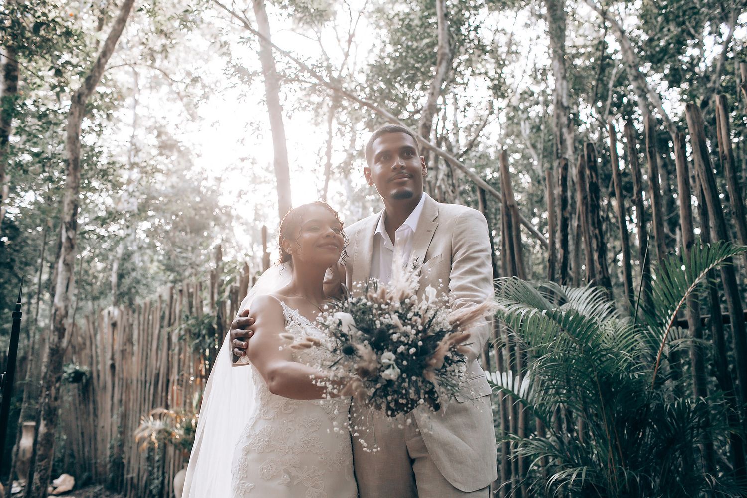 Newlyweds embrace tenderly in a sun-drenched forest with bamboo fencing in the background.