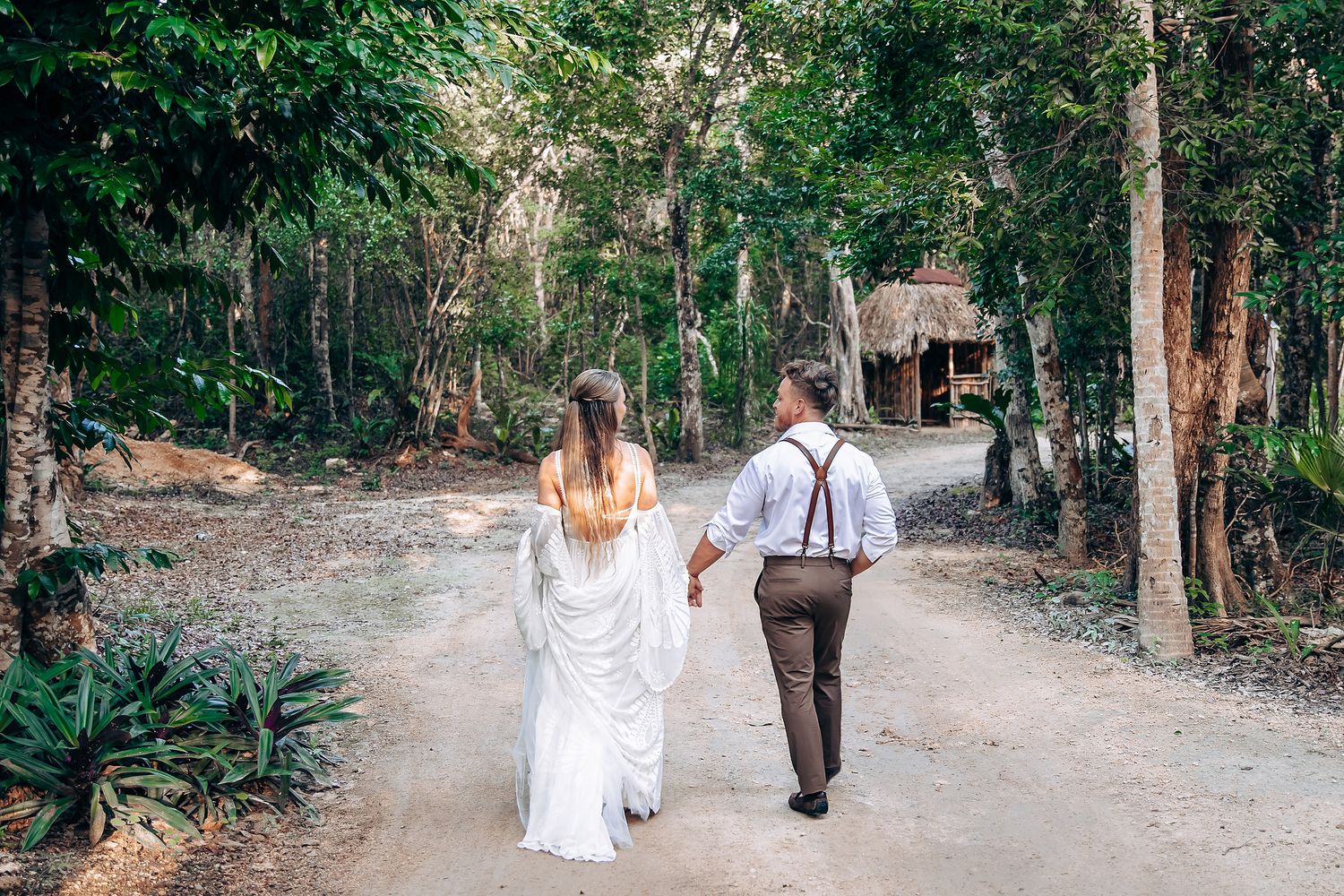 A couple walks down a tree-lined path towards a rustic wooden structure in a tropical forest setting.
