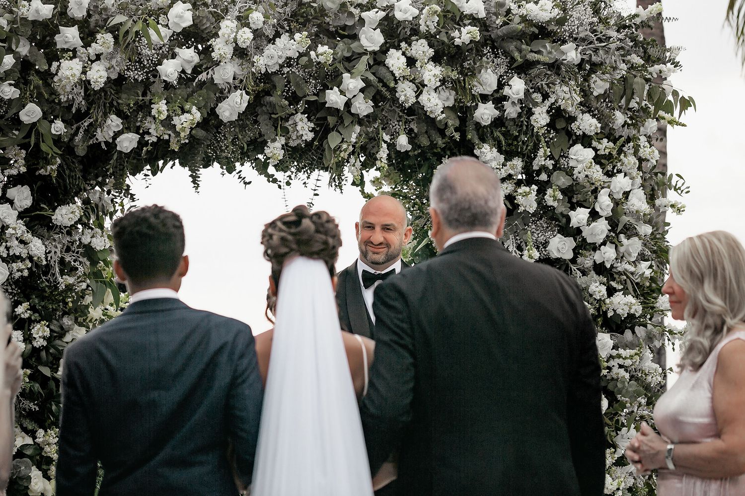 Wedding guests gather under an elegant white floral arch during an outdoor ceremony.