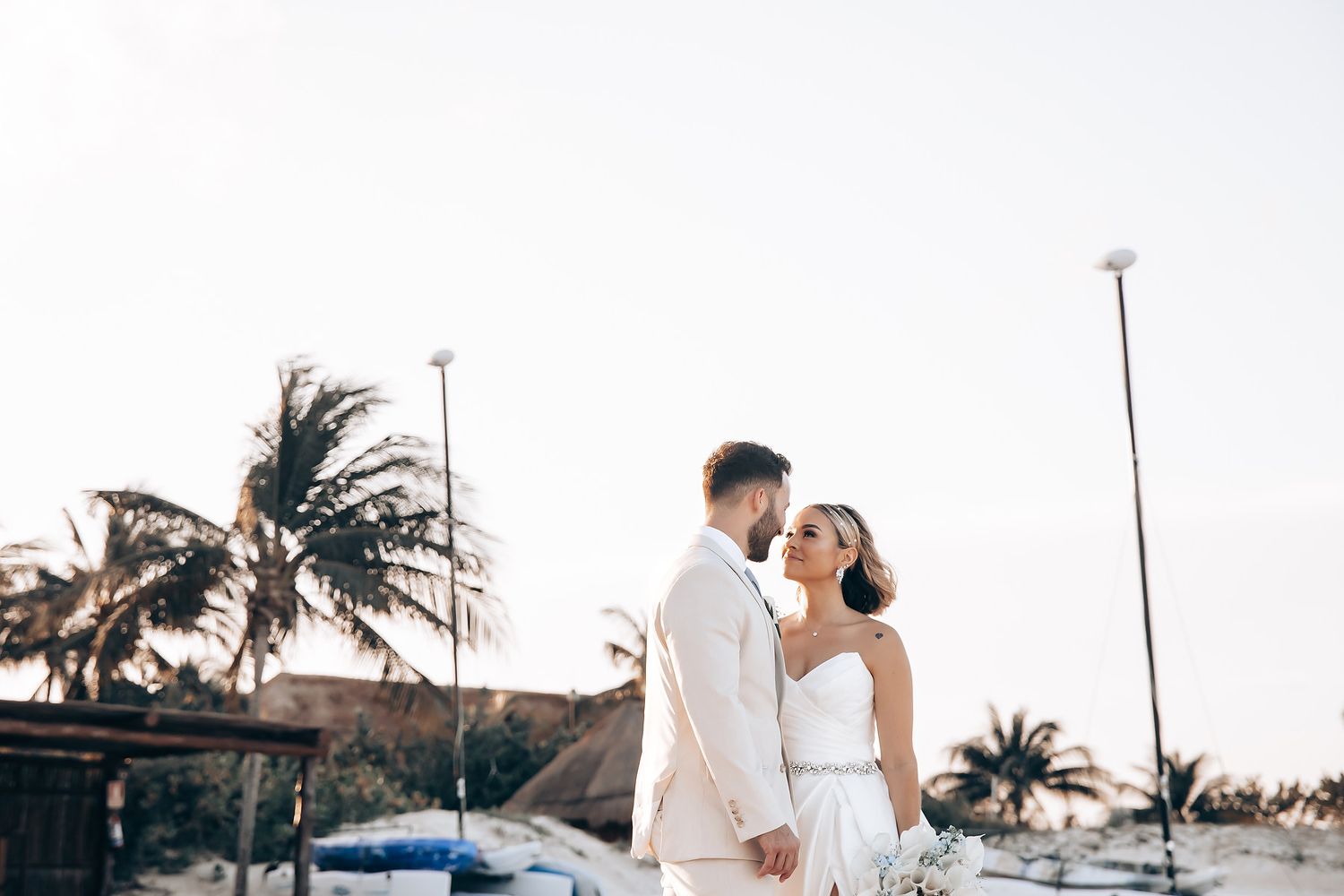 Beachfront wedding ceremony setup with white chairs and tropical palm trees at sunset.