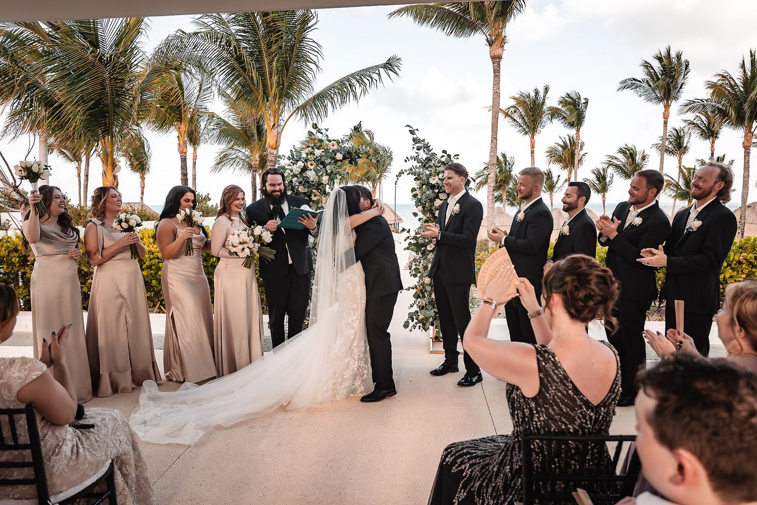 Wedding guests gather under swaying palm trees at an elegant outdoor beach ceremony.