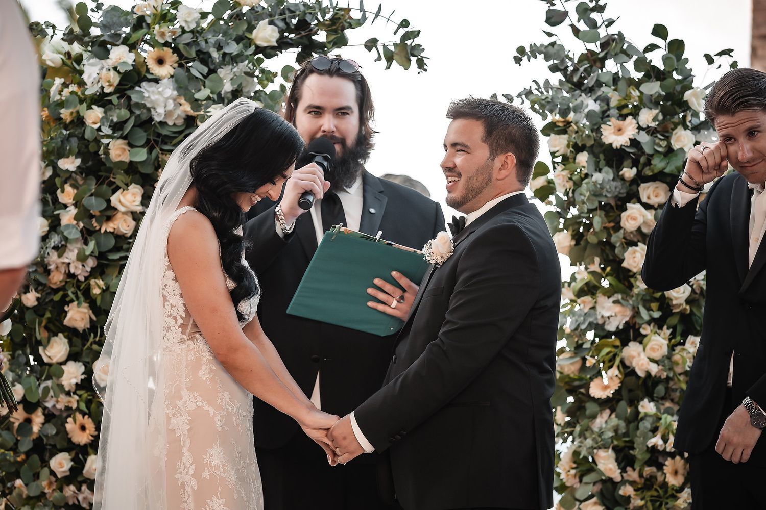 Wedding ceremony in front of floral arch with white roses and greenery.