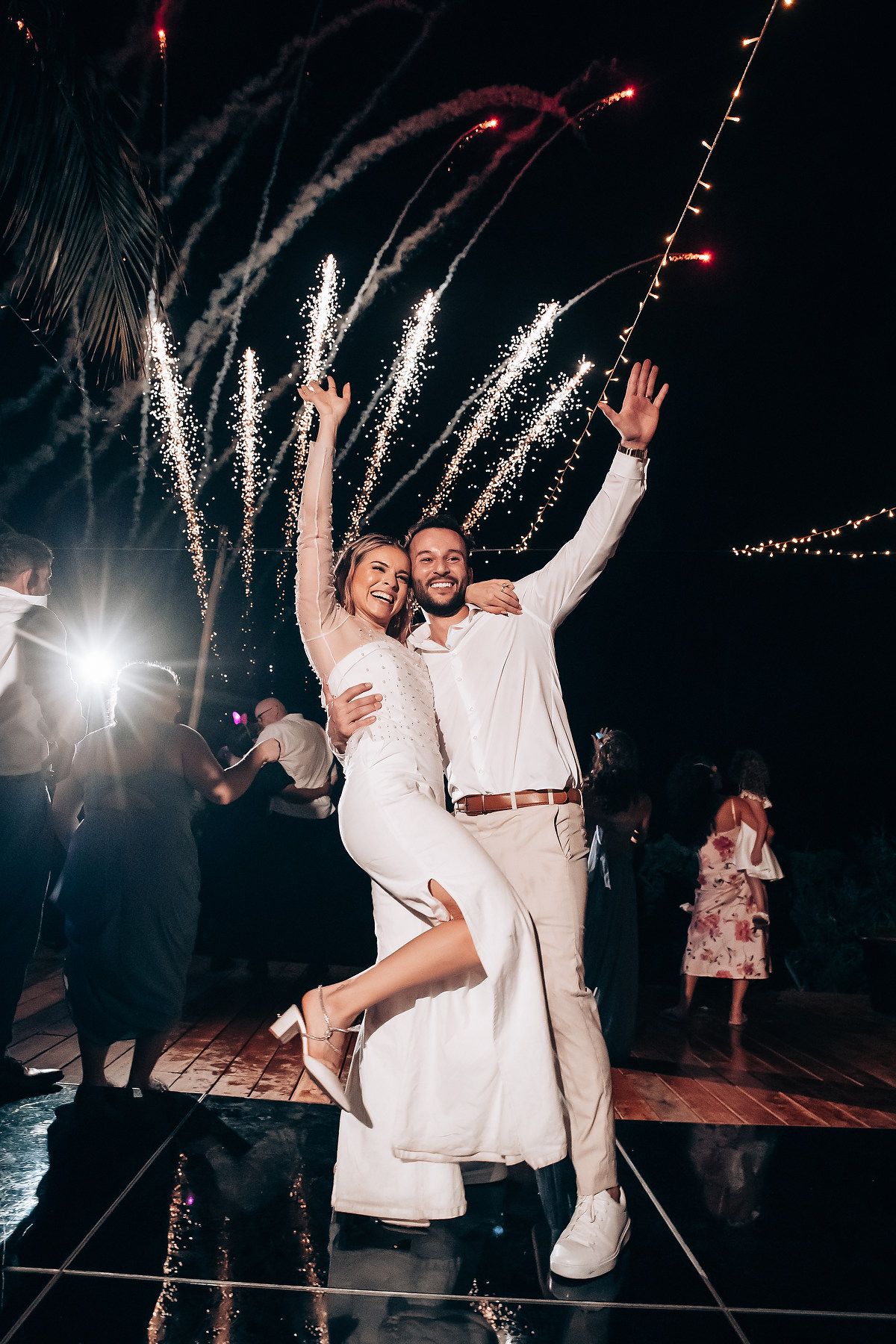 Couple celebrating on dance floor with firework sparklers in background at night.