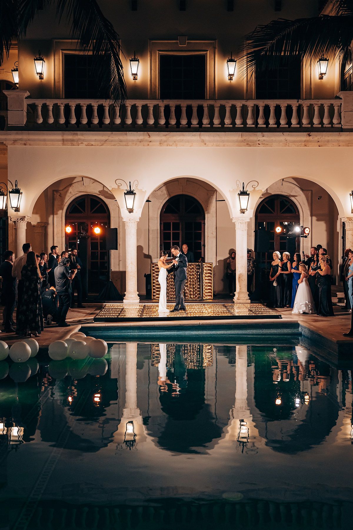Elegant nighttime wedding reception by an illuminated pool with arched columns and reflective water.