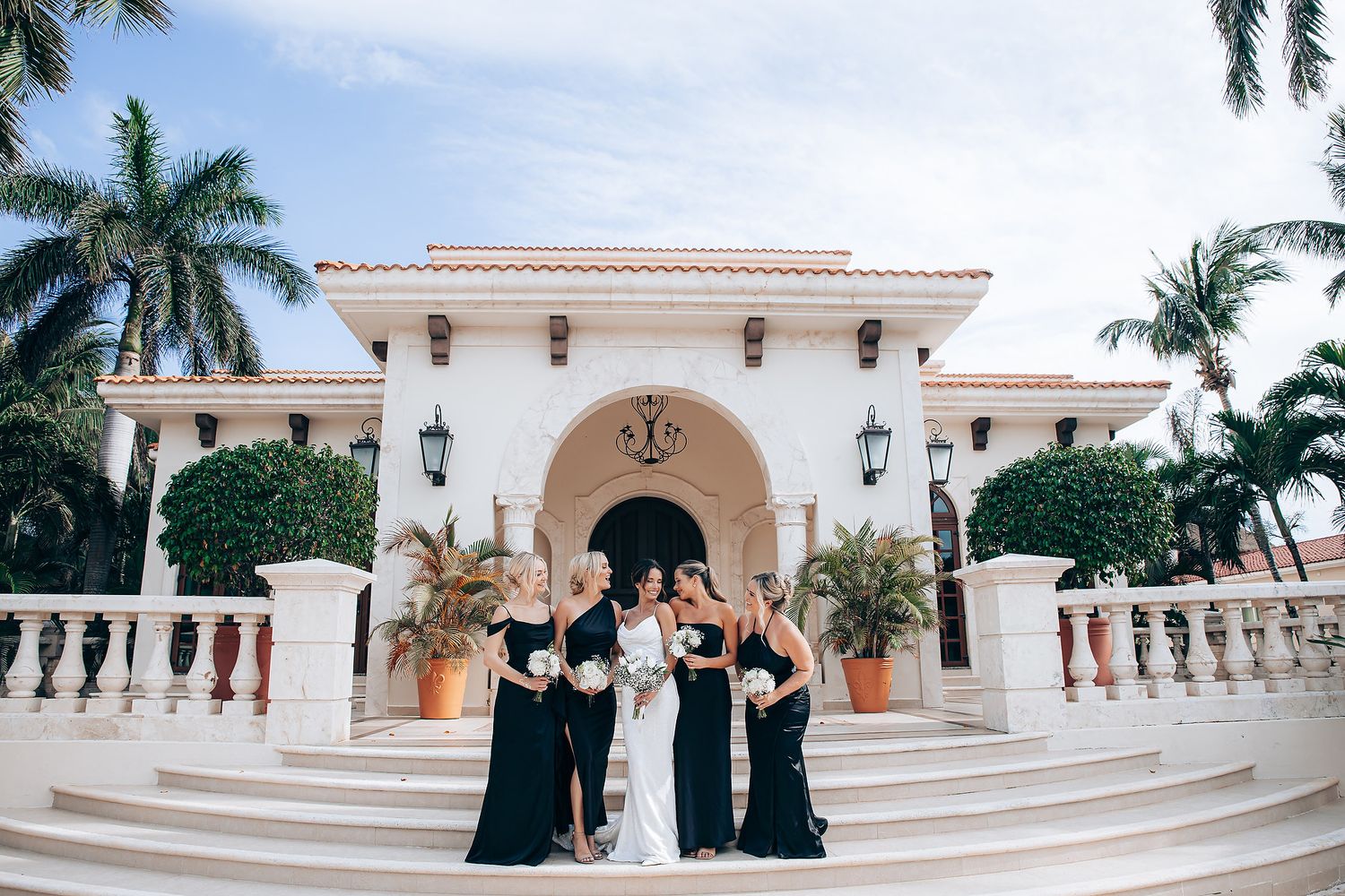 A wedding party poses on grand white marble steps in front of a Mediterranean-style mansion with palm trees.