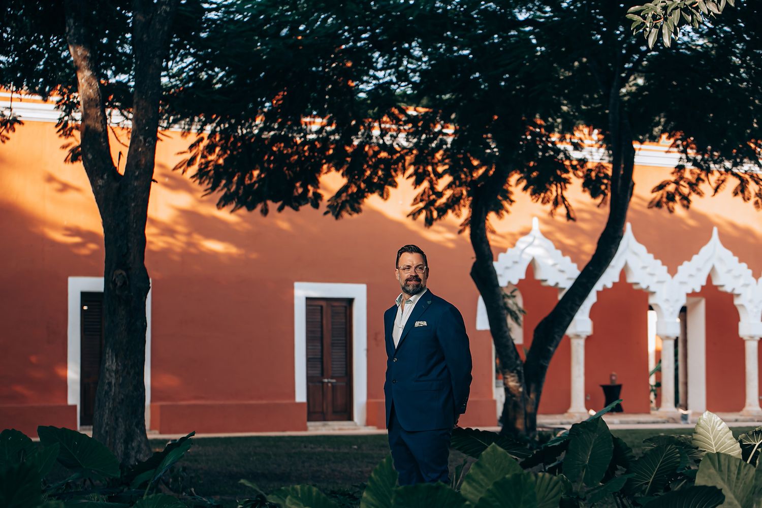 Wedding party poses under tall trees at sunset with a rustic red building in the background.