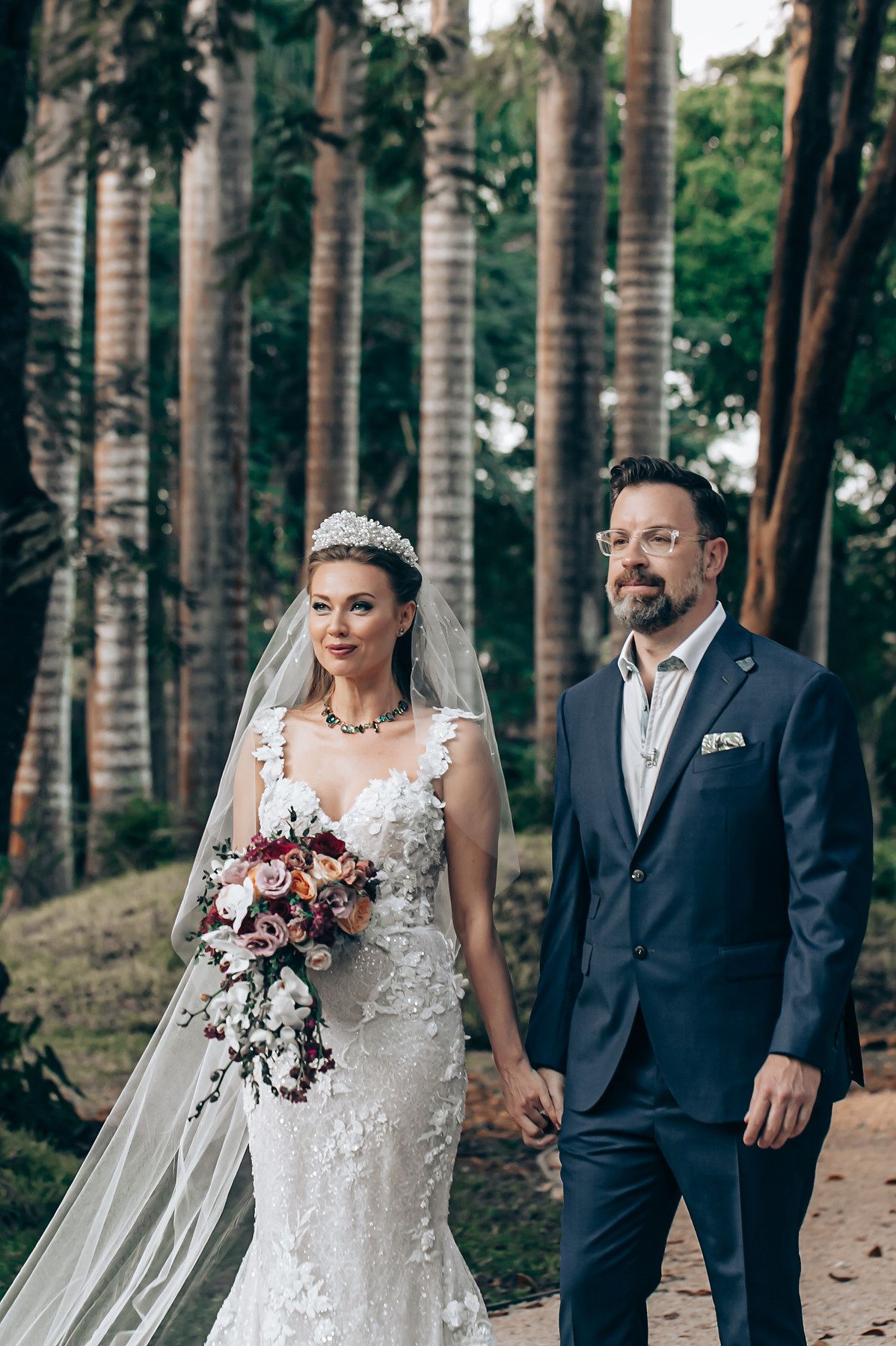 A wedding couple stands together on a path lined with tall palm trees while holding a colorful bouquet.