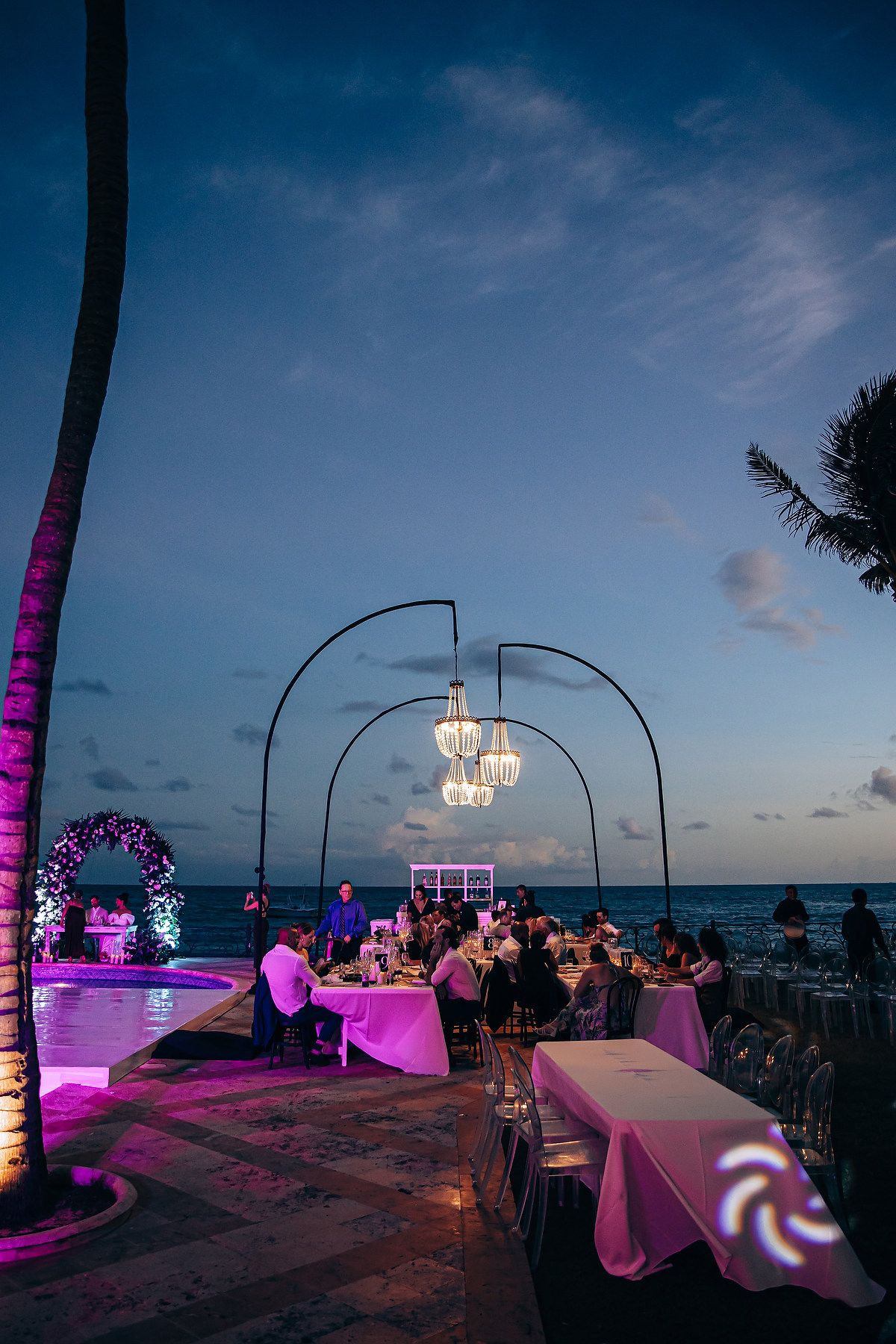 Elegant beachside wedding reception setup with purple lighting, chandeliers and palm trees against a twilight ocean view.