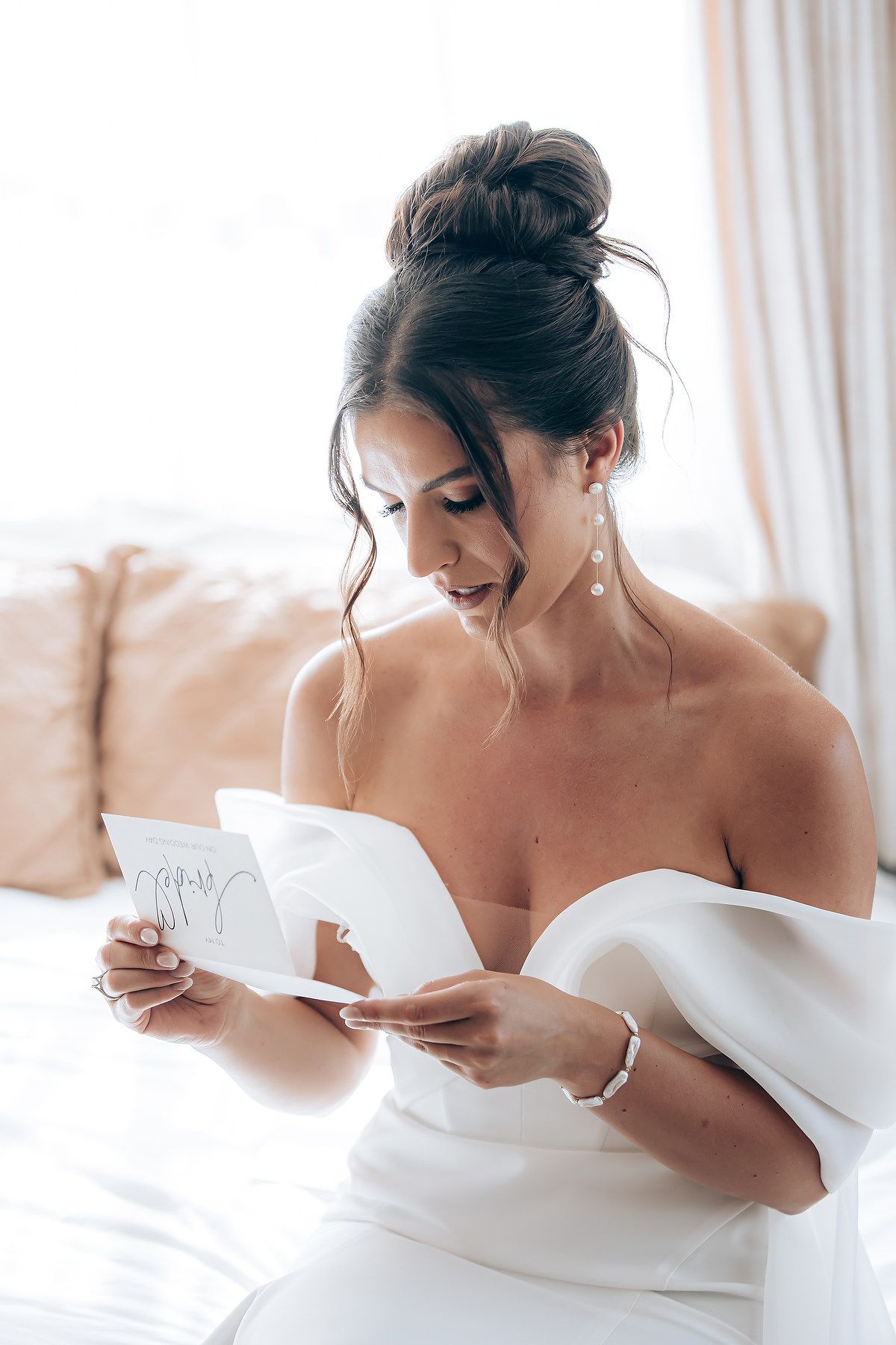 A person in a white off-shoulder dress reads a letter while sitting on a couch with soft lighting and curtains.