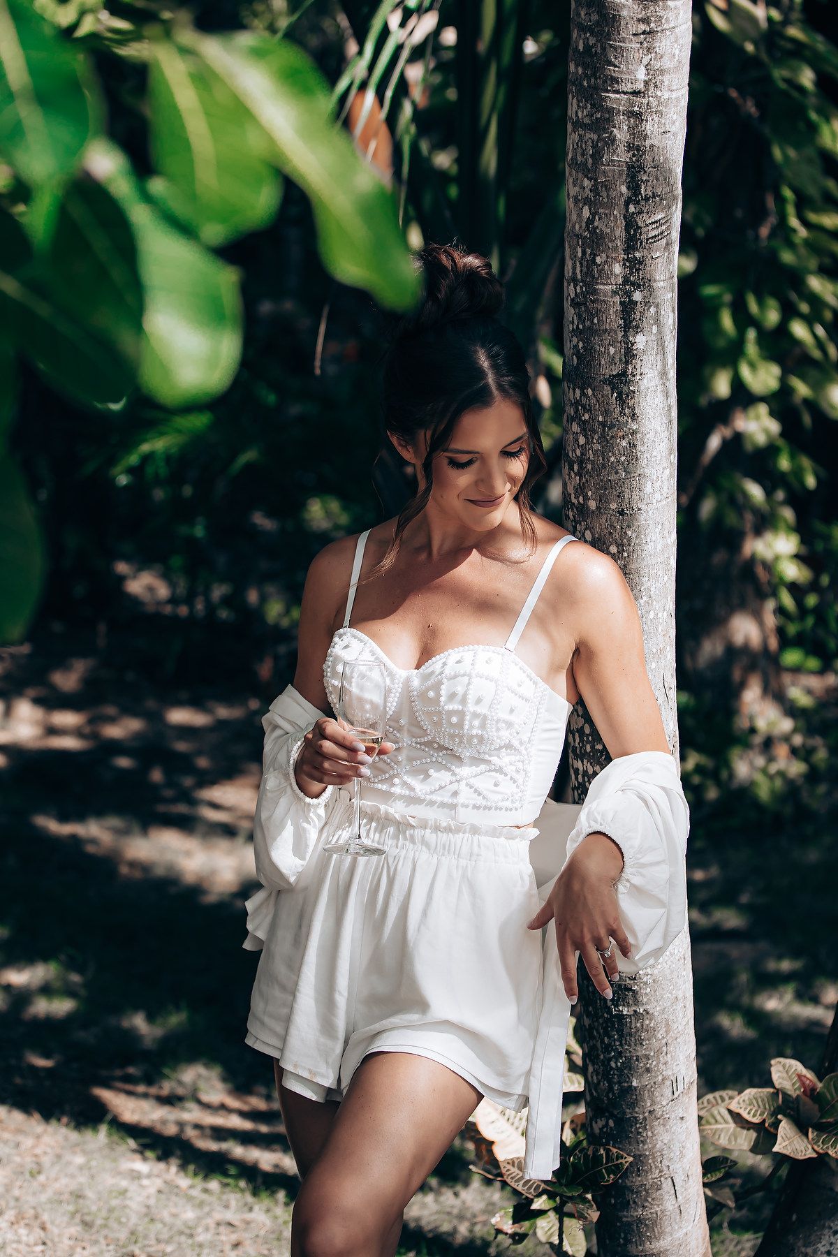 A woman in a white lace romper poses against a tree trunk in a natural outdoor setting with green foliage.