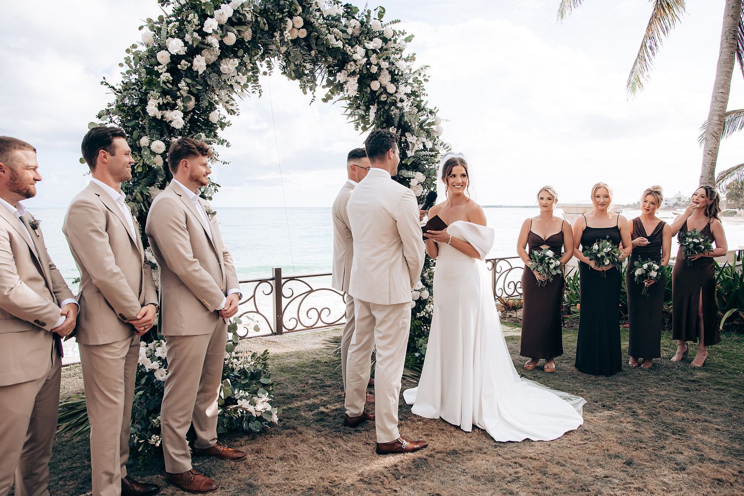 Wedding party stands in line on a tropical beach with palm trees and ocean in the background during sunset ceremony.