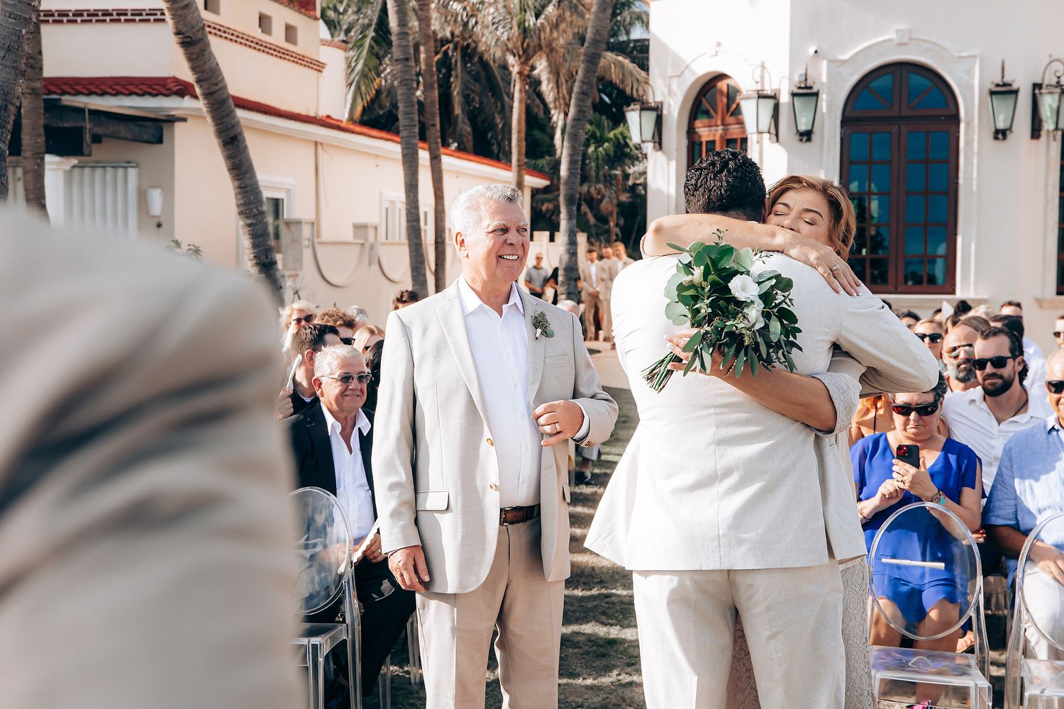 A wedding celebration scene with people in formal attire gathered outside a white Mediterranean-style building.