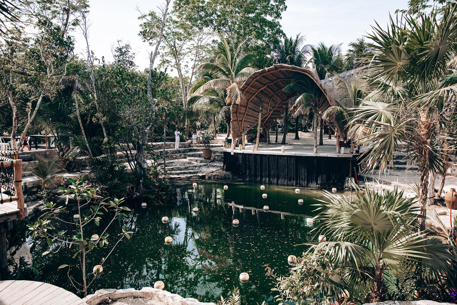 A wooden gazebo overlooks a tropical cenote pool surrounded by lush palm trees and vegetation in Mexico.