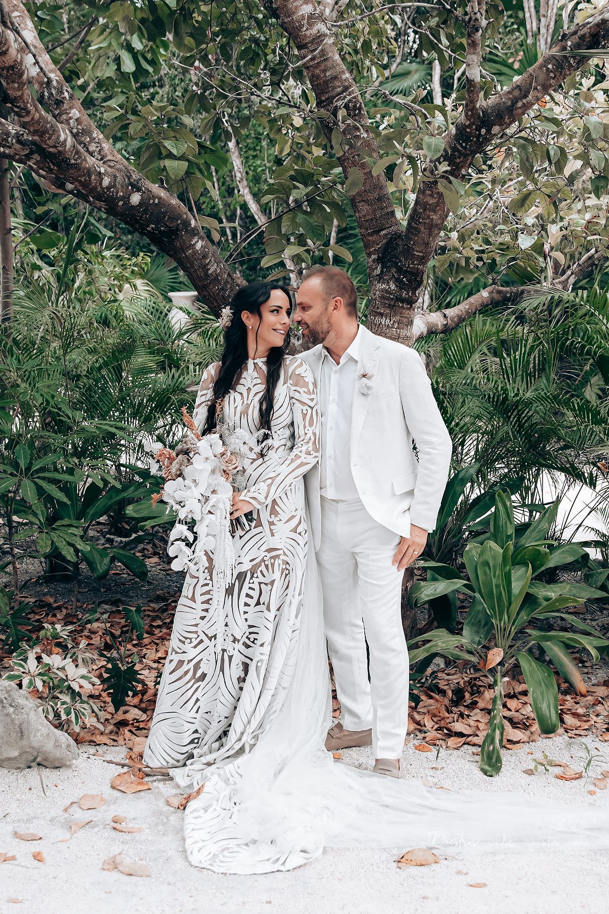 A couple in white attire poses together in a romantic outdoor garden setting with tropical foliage.