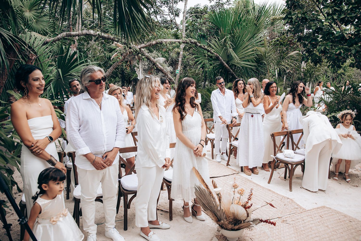 Wedding guests dressed in all white gather in a tropical garden setting with palm trees in the background.
