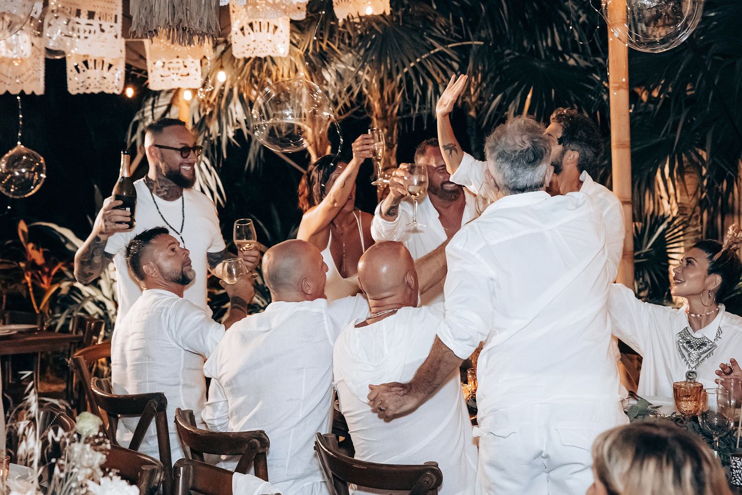 People in white clothing dance and celebrate together at a festive party with string lights overhead.