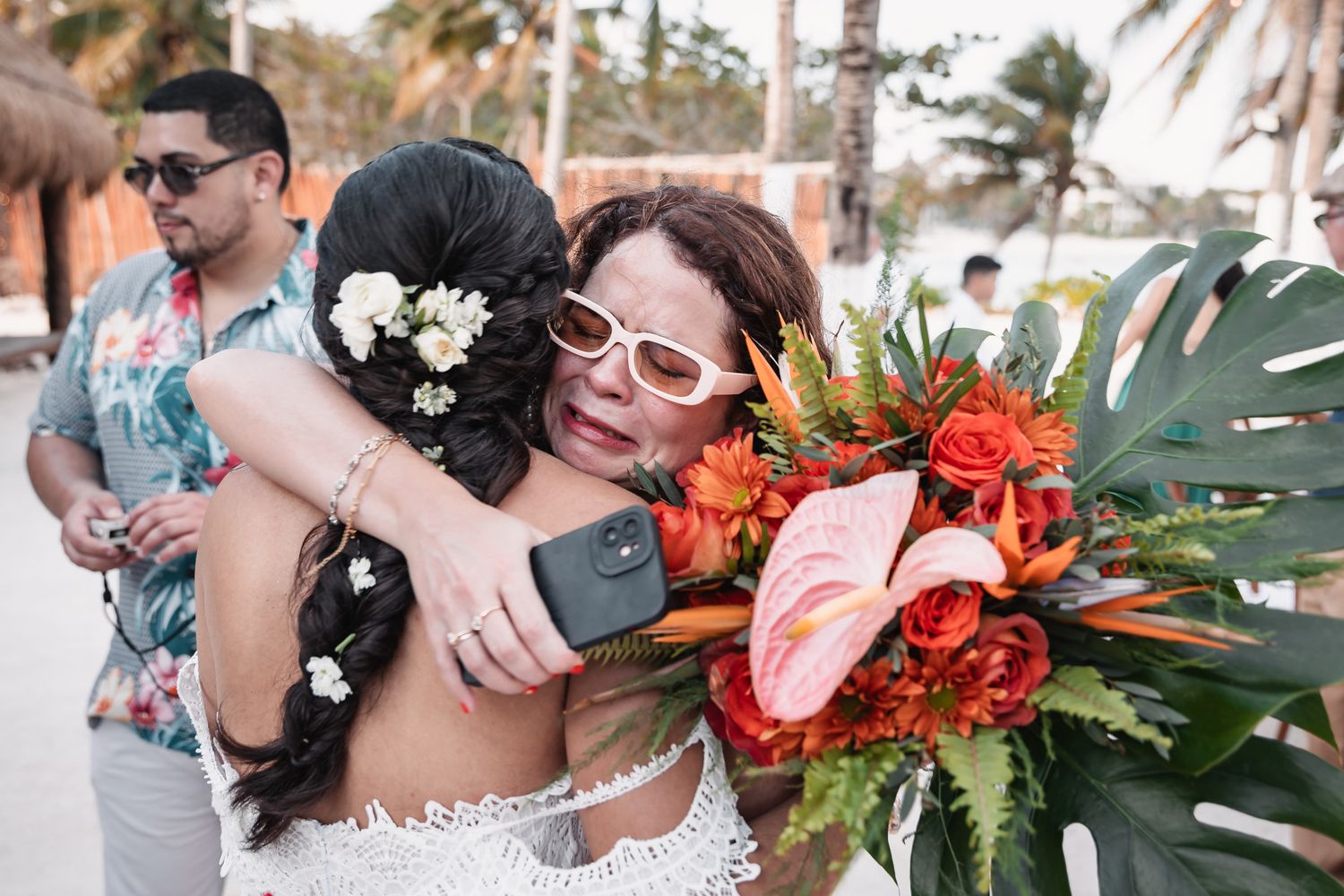 Bride hugging guest while holding vibrant tropical wedding bouquet on beach.