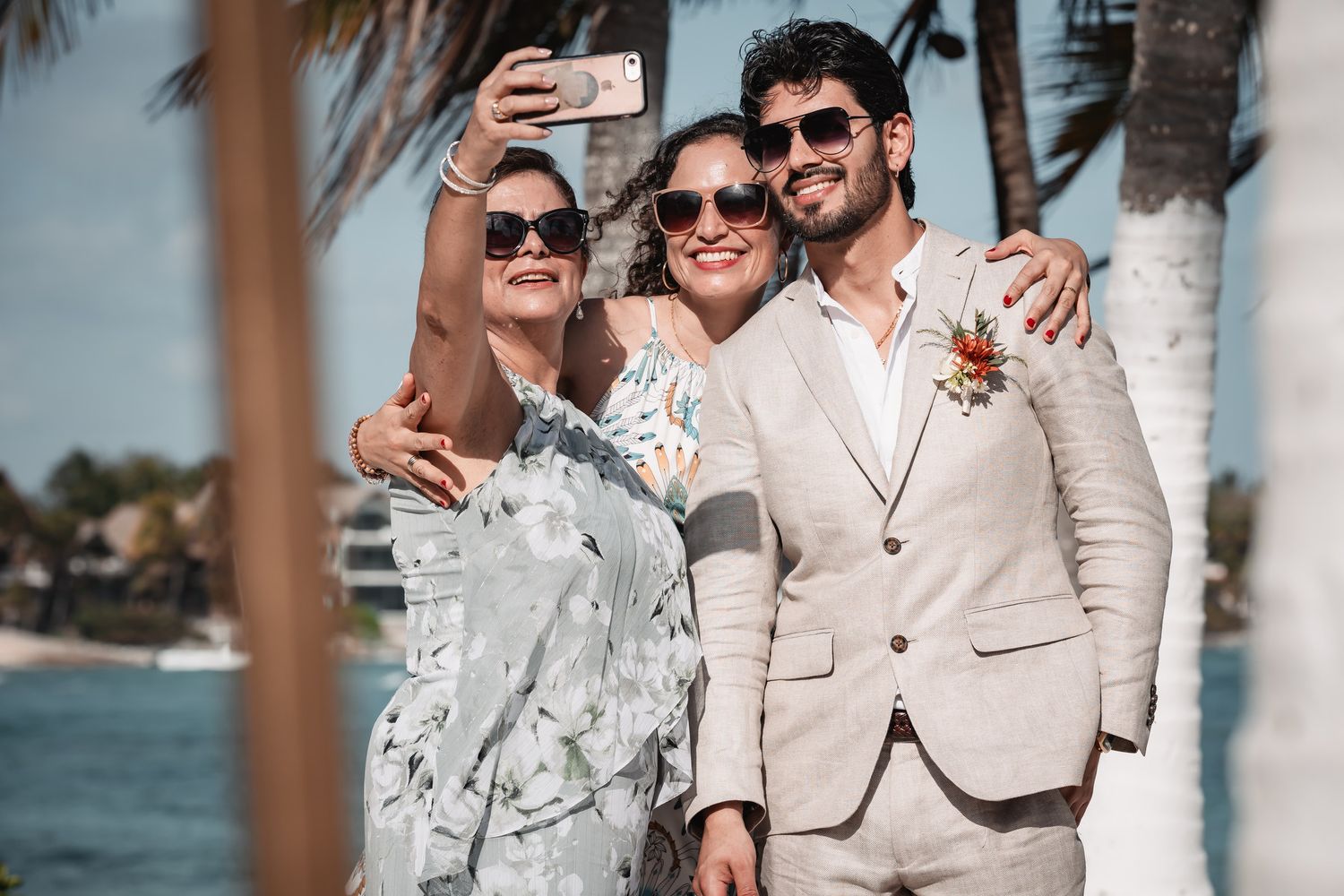 Groom in beige suit taking selfie with two women at tropical beach wedding.