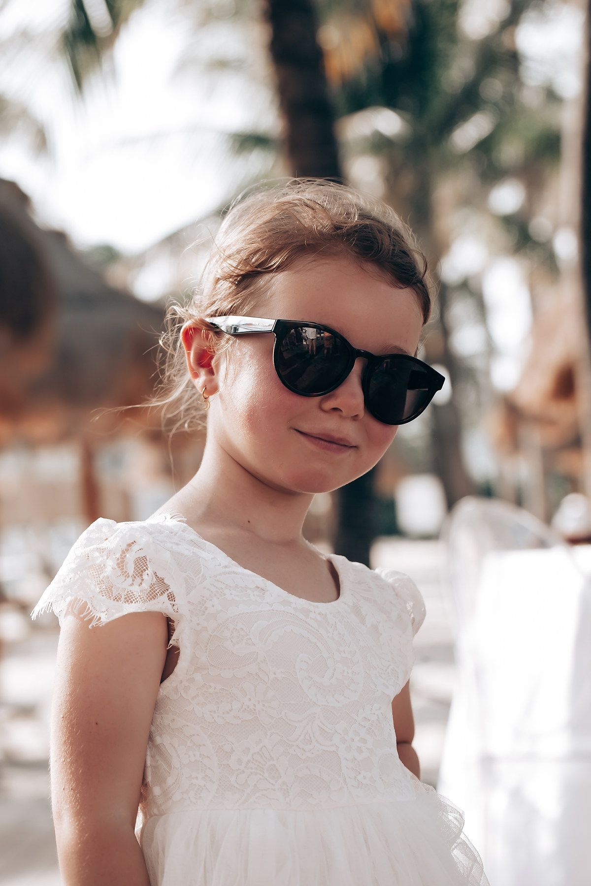 Young attendee in white lace dress wearing sunglasses at tropical event.