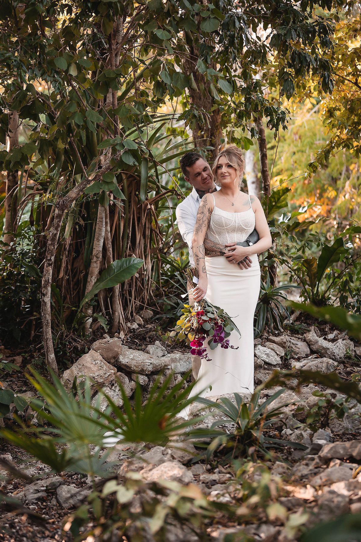Bride and groom embrace in a lush tropical garden, bride holding a vibrant bouquet in a white fitted gown.