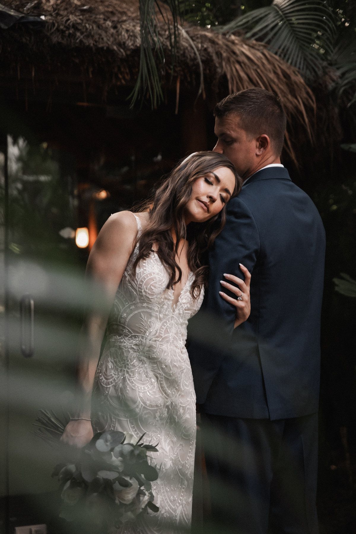 Romantic portrait of bride in lace gown and groom in navy suit embracing in lush tropical garden.