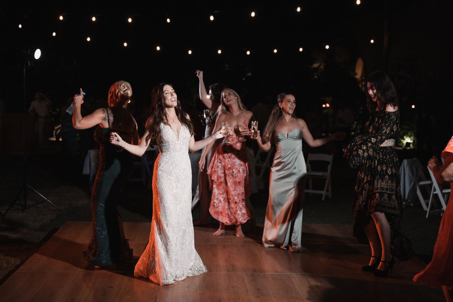 Bride and bridesmaids dancing joyfully at a wedding reception under warm string lights.