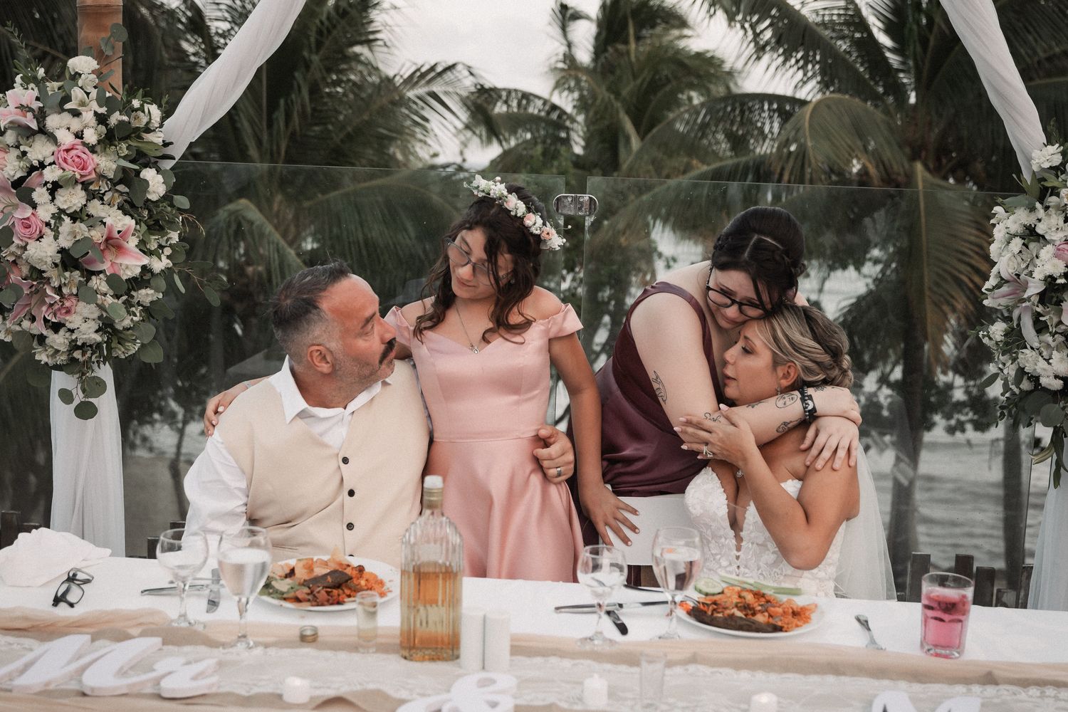 Bride, groom, and two bridesmaids share an emotional group hug at decorated beachside wedding reception table.
