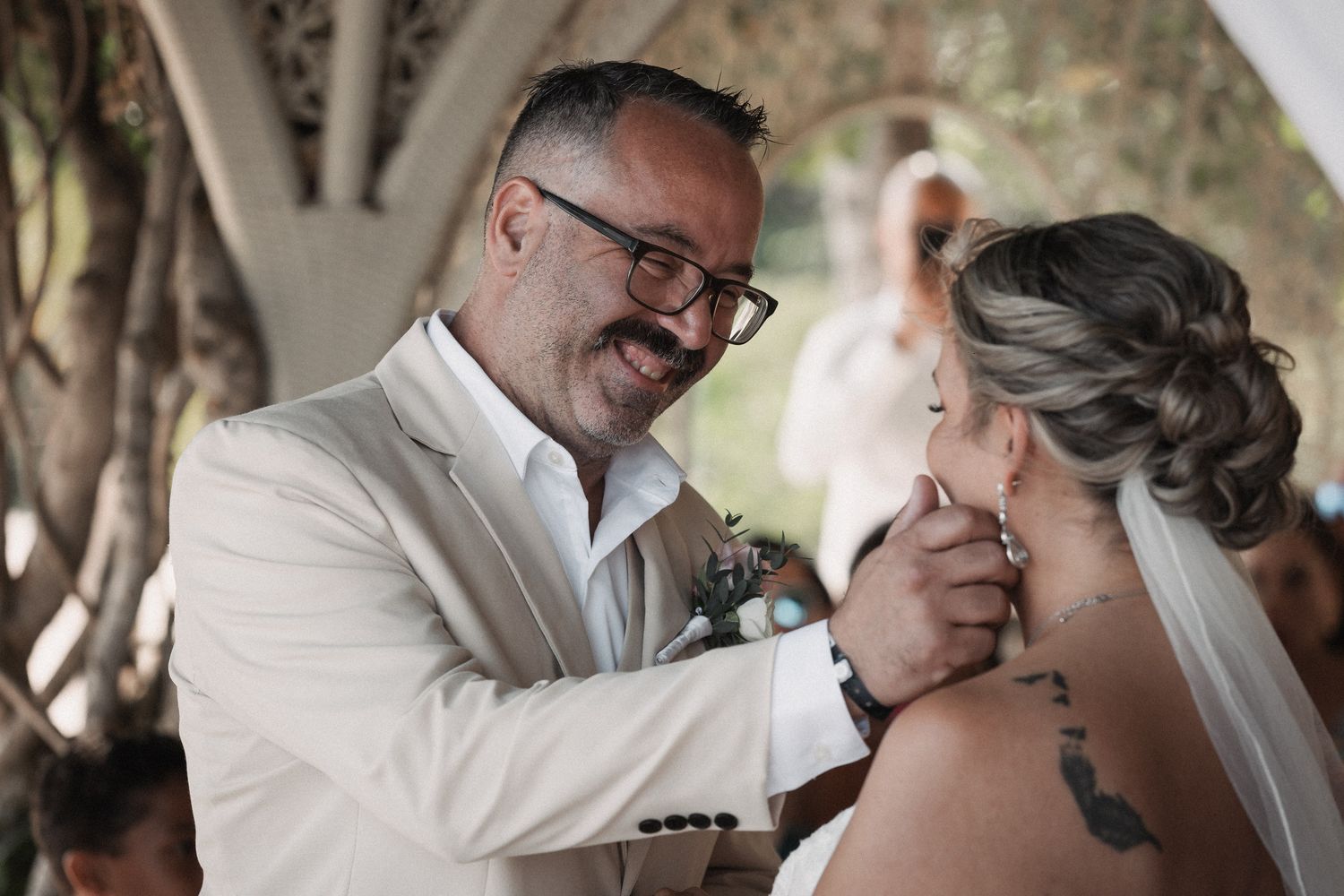 Groom in beige suit tenderly touches brides face during outdoor wedding ceremony.