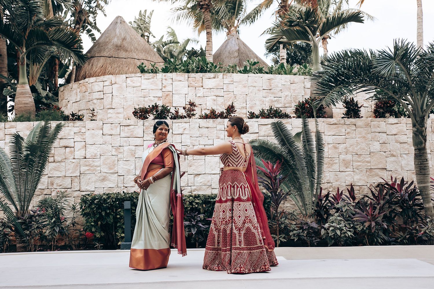 Two women in traditional Indian wedding attire stand against a stone wall surrounded by tropical palm trees.