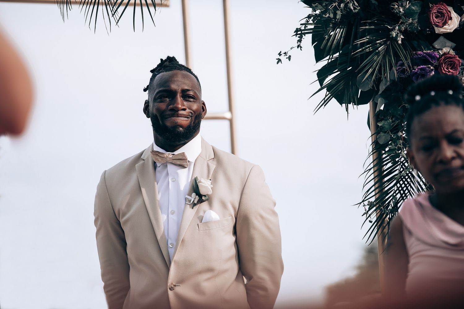Groom in light beige suit with bowtie stands among tropical decor.
