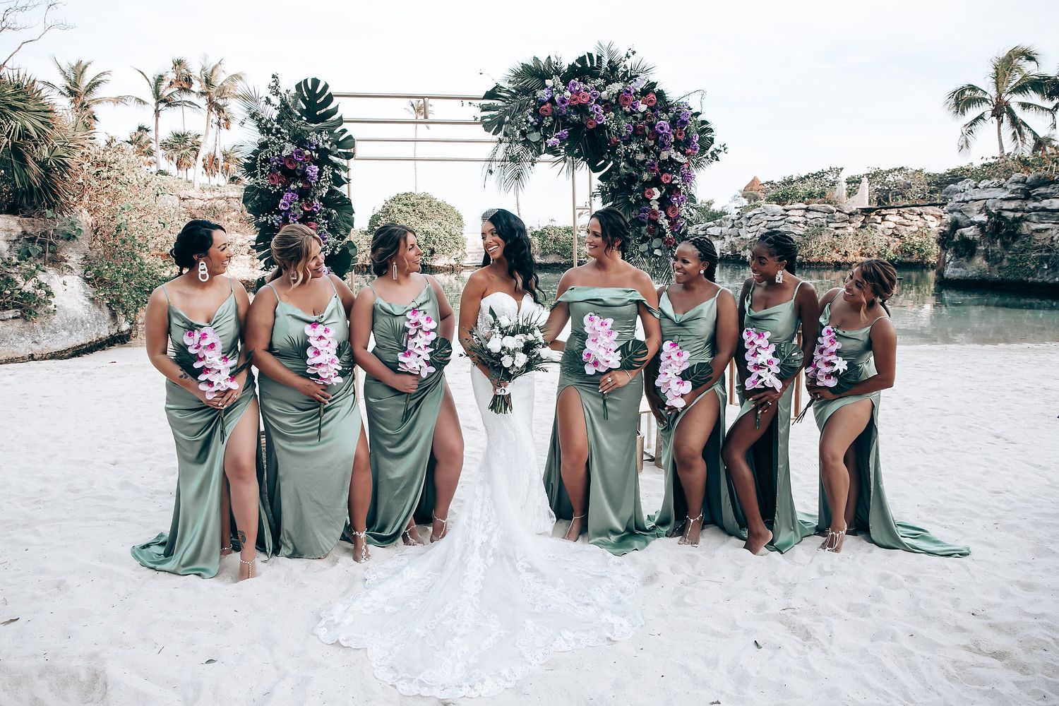 Bridesmaids in sage green dresses pose together on a beach with floral archway.