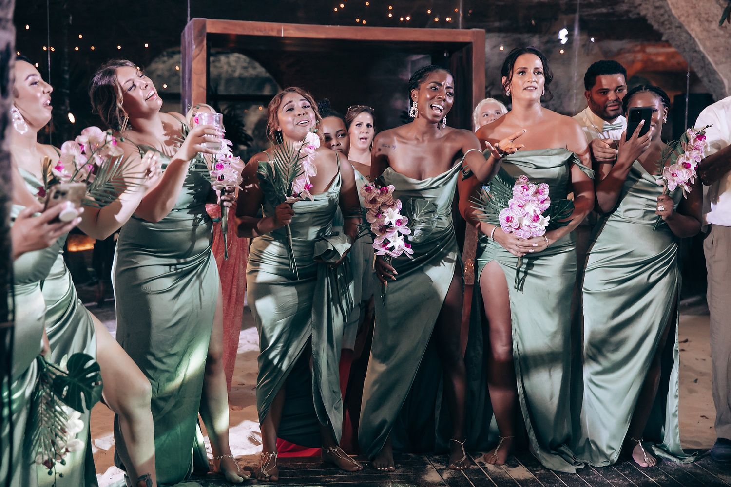 Wedding party in sage green dresses poses together at reception venue.