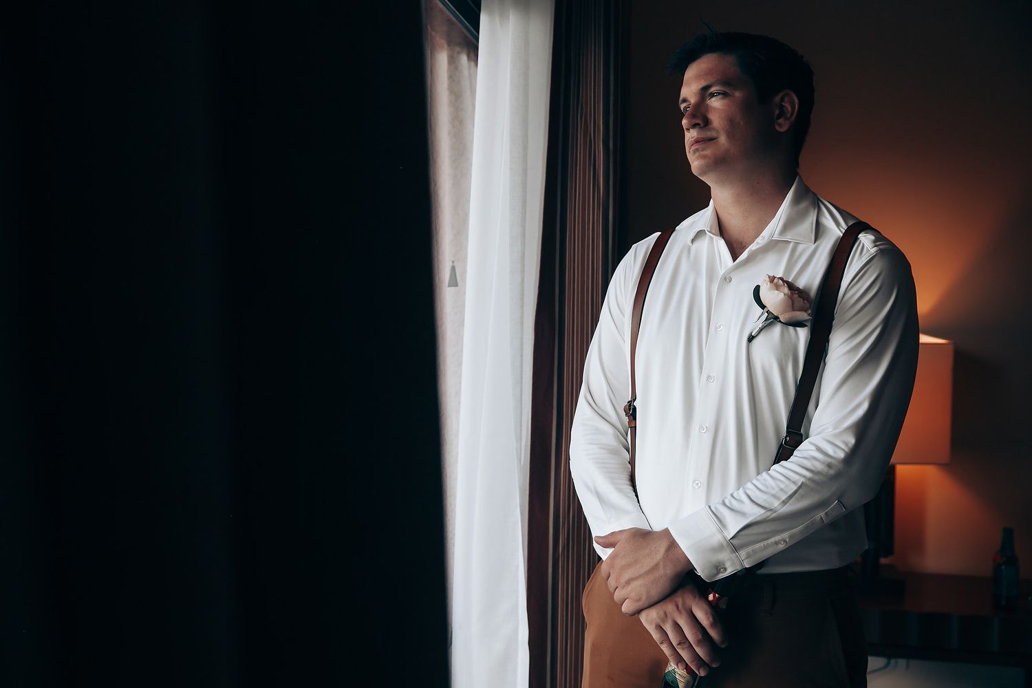 Groom and groomsmen getting ready in formal attire by a window with dramatic lighting.