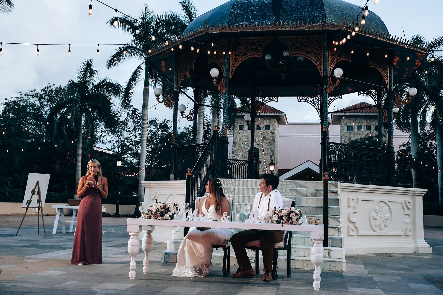 Outdoor wedding reception setup with string lights and palm trees at tropical venue.