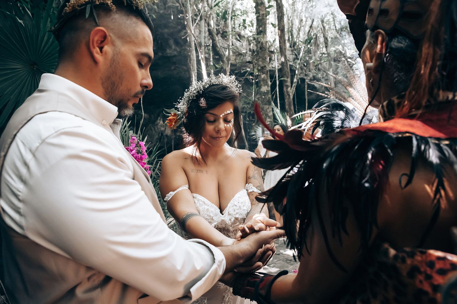 A couple participates in a traditional ceremony under a forest canopy with vibrant pink flowers.