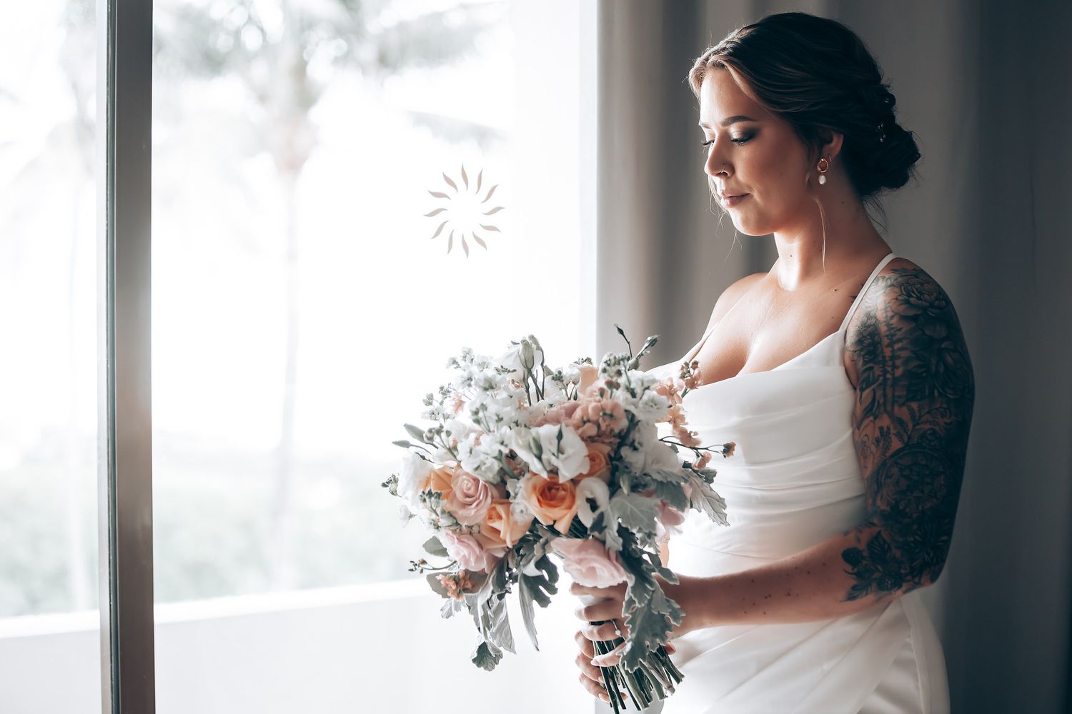 A bride in a white wedding dress holds a romantic blush and white floral bouquet near a window.