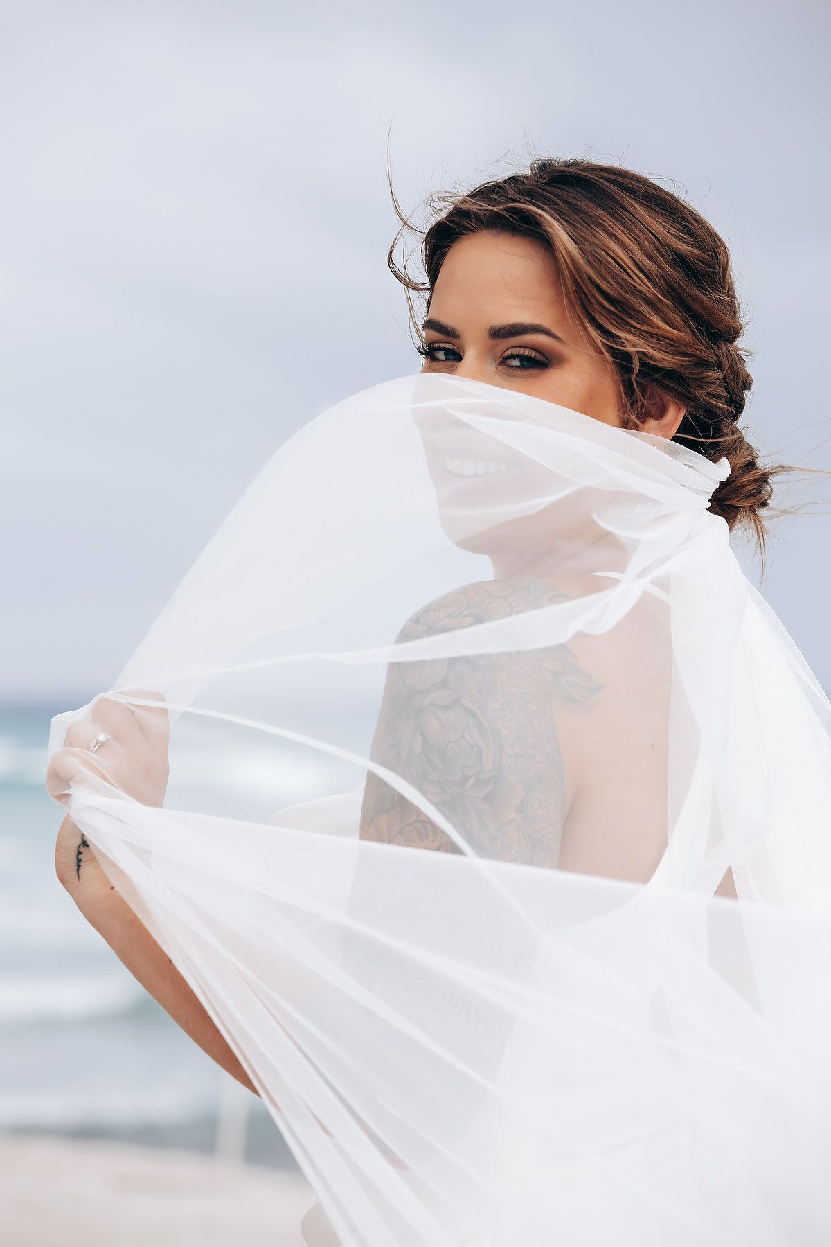 A delicate white veil flows in the wind against a cloudy beach backdrop.