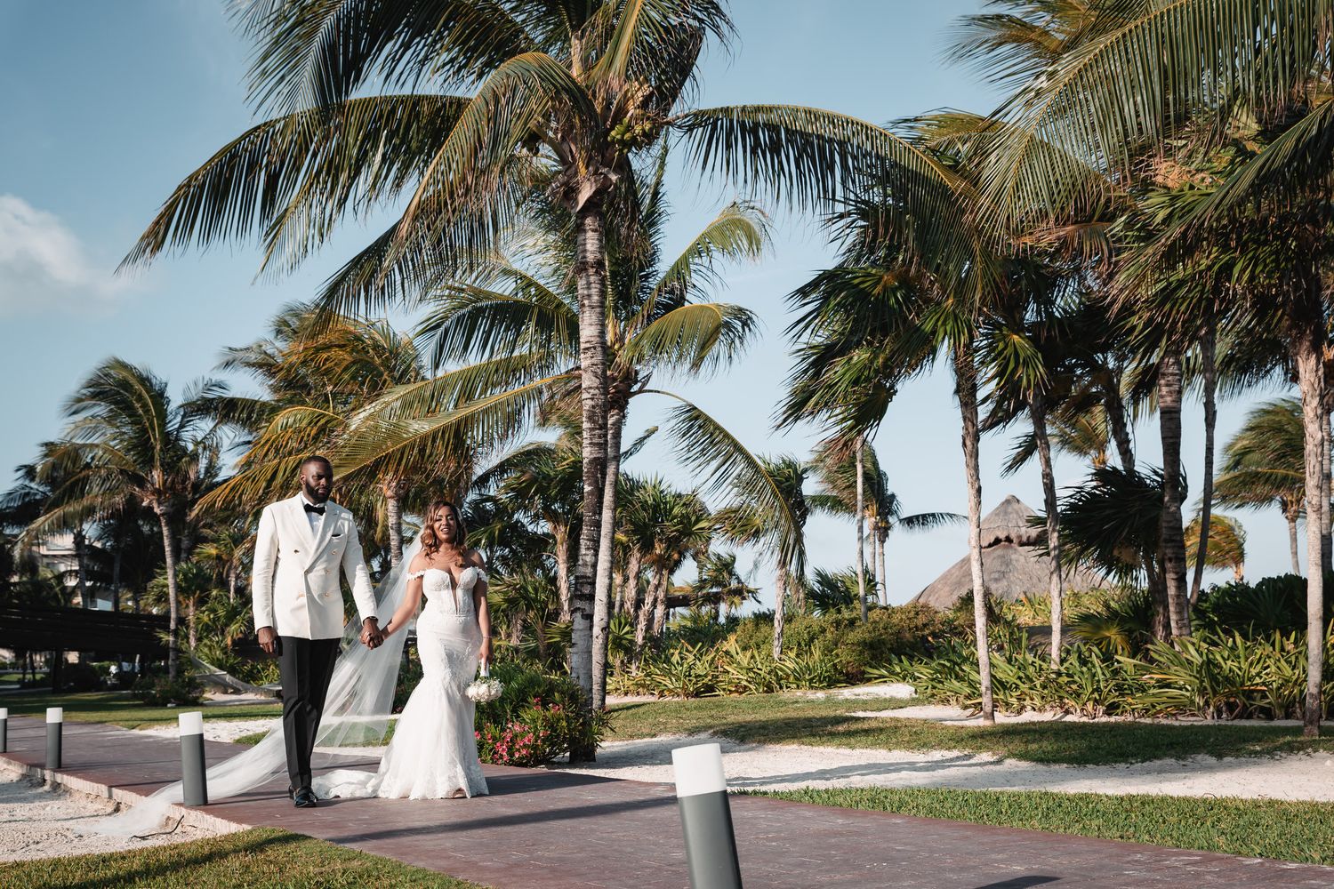 Newlywed couple walking hand in hand on a tropical resort pathway surrounded by lush palm trees on their wedding day.