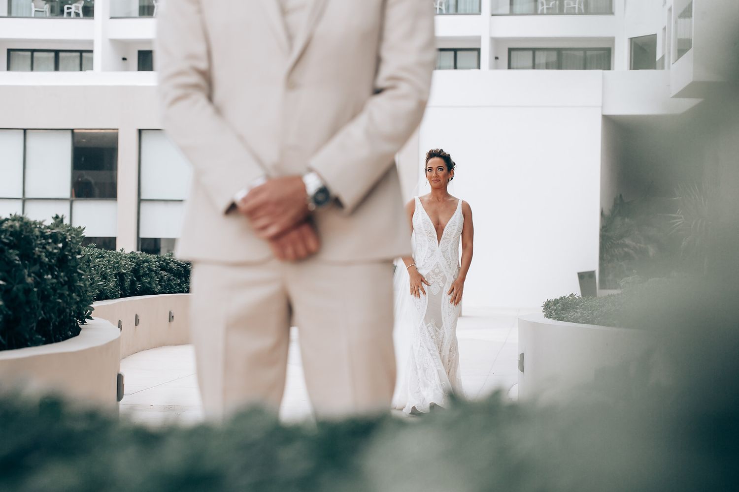A bride in a white gown stands on a balcony while someone in a beige suit is seen in the foreground.