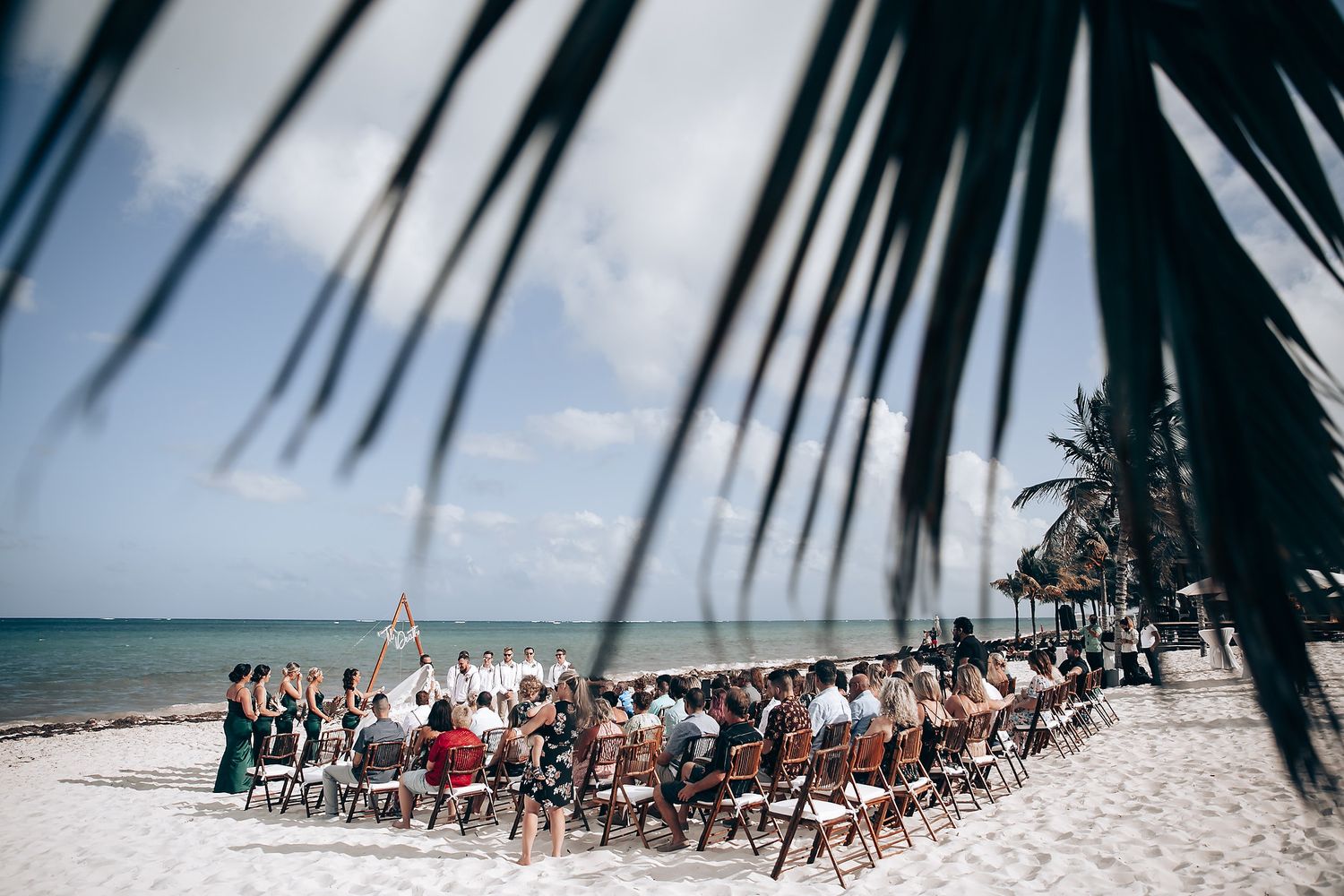 Beach wedding ceremony setup with white chairs and palm fronds framing a tropical oceanfront view.