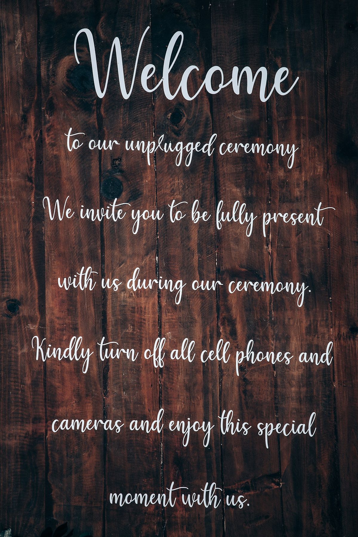 Rustic wooden wall sign welcoming guests to an unplugged ceremony with elegant white script lettering.