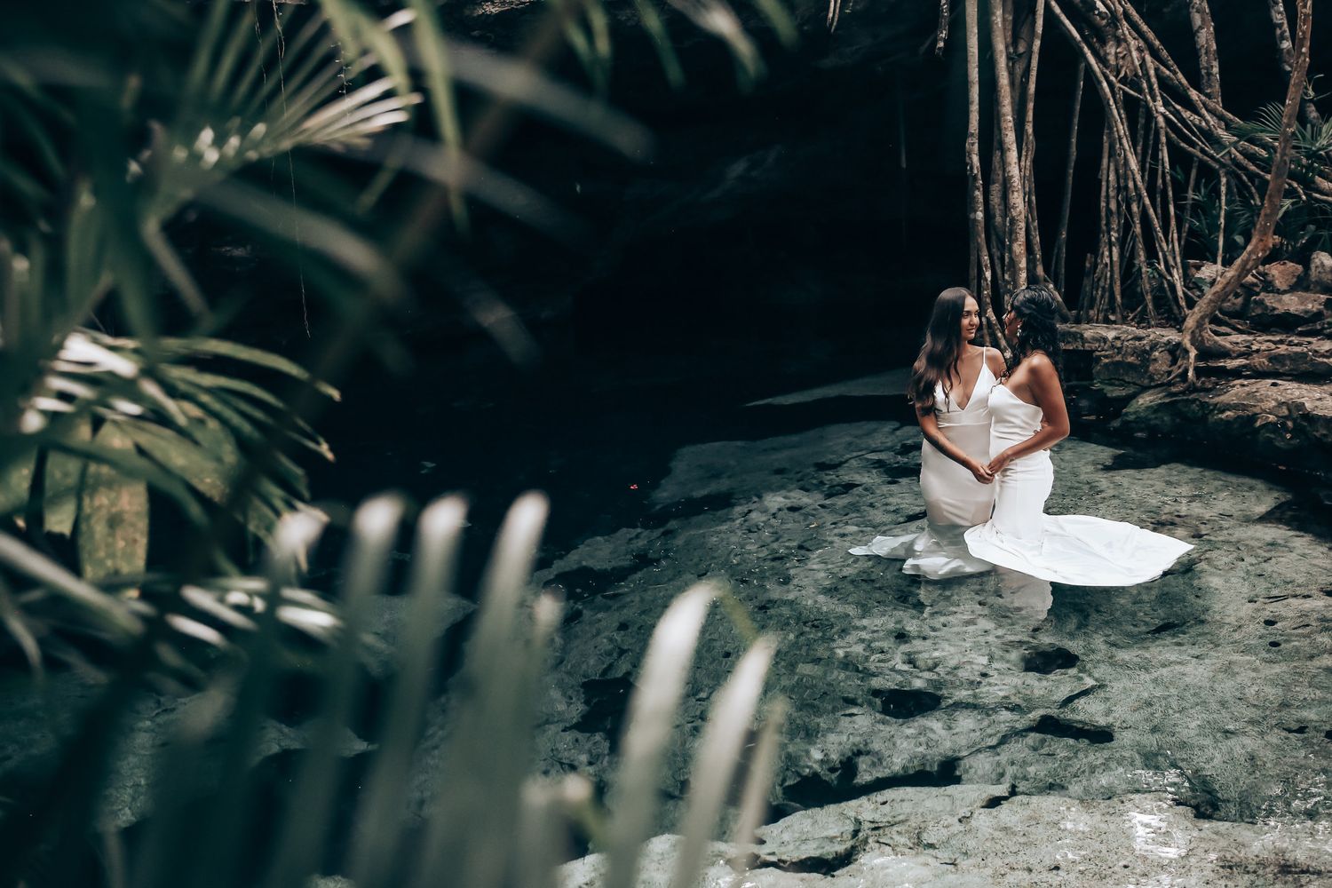 Artistic wedding photos captured through tropical leaves show two people in white dresses.
