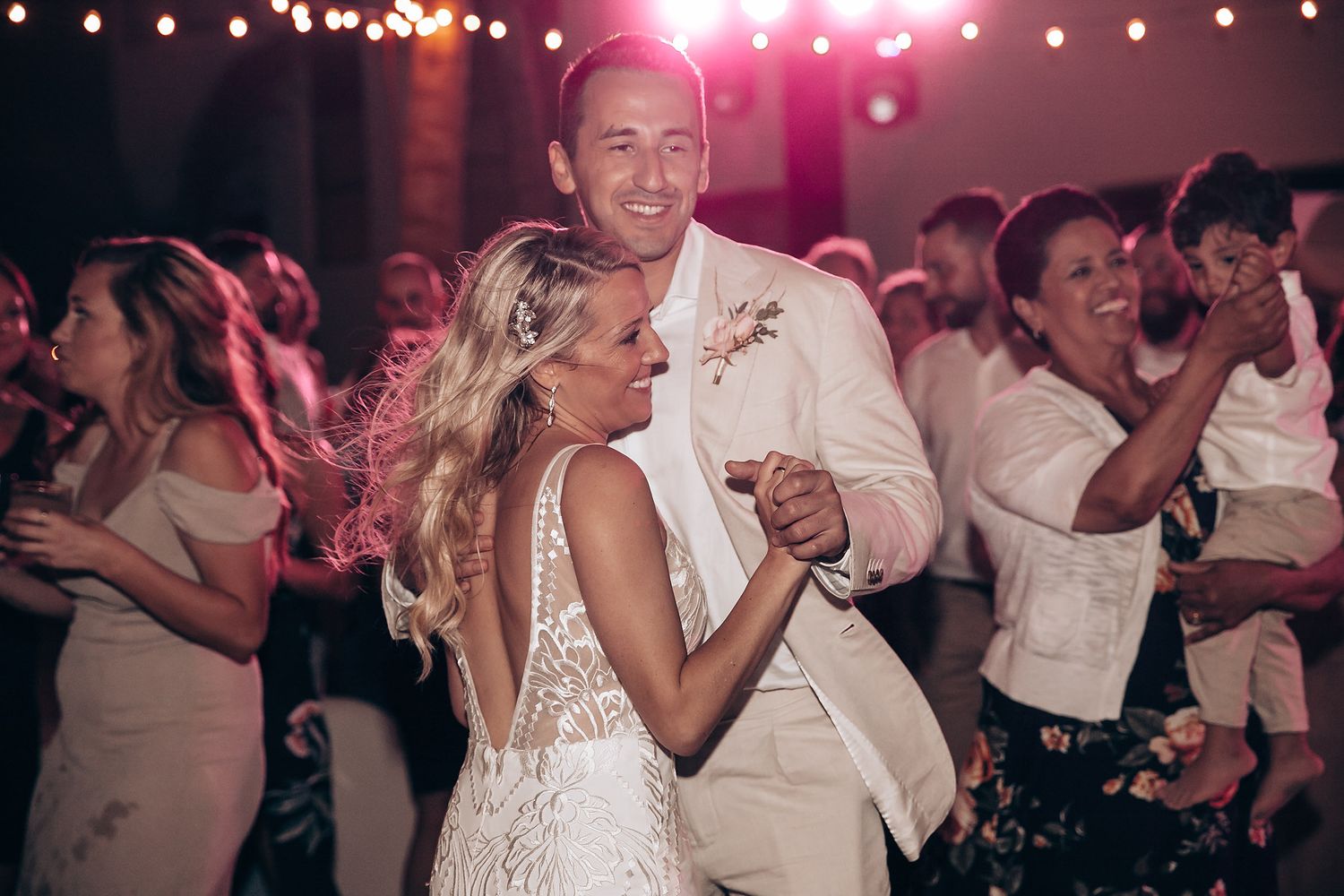 Wedding party dancing under string lights at evening reception.