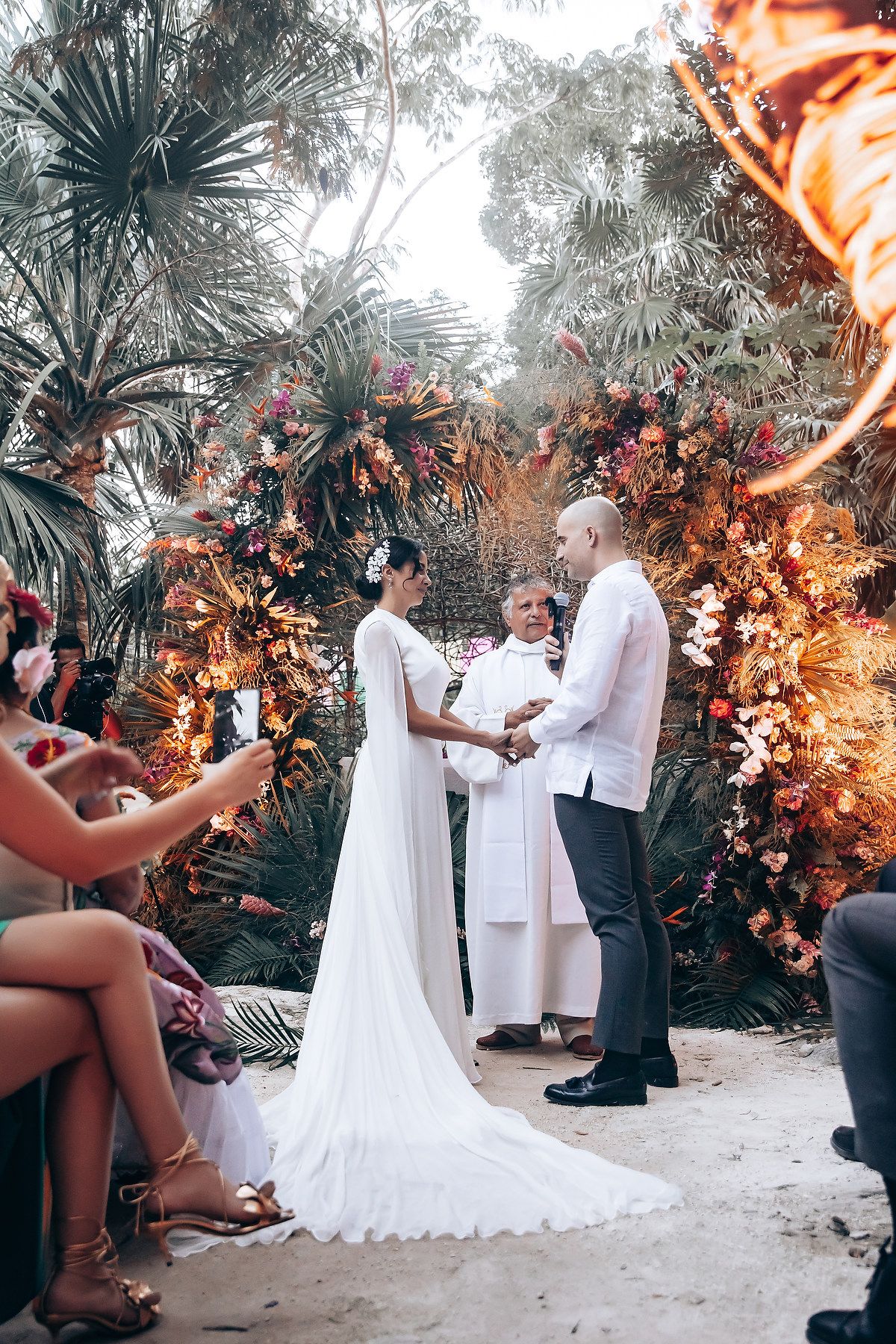 A wedding ceremony takes place under illuminated floral arches in a tropical garden setting.