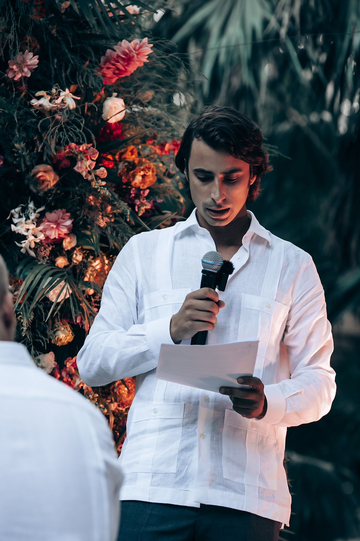 Wedding officiant reads ceremony text surrounded by coral flowers and tropical foliage in an outdoor setting.