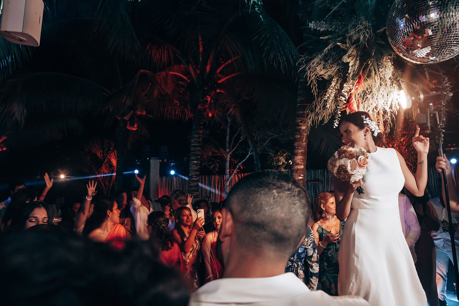 Wedding reception dance party under palm trees with atmospheric lighting and a disco ball.