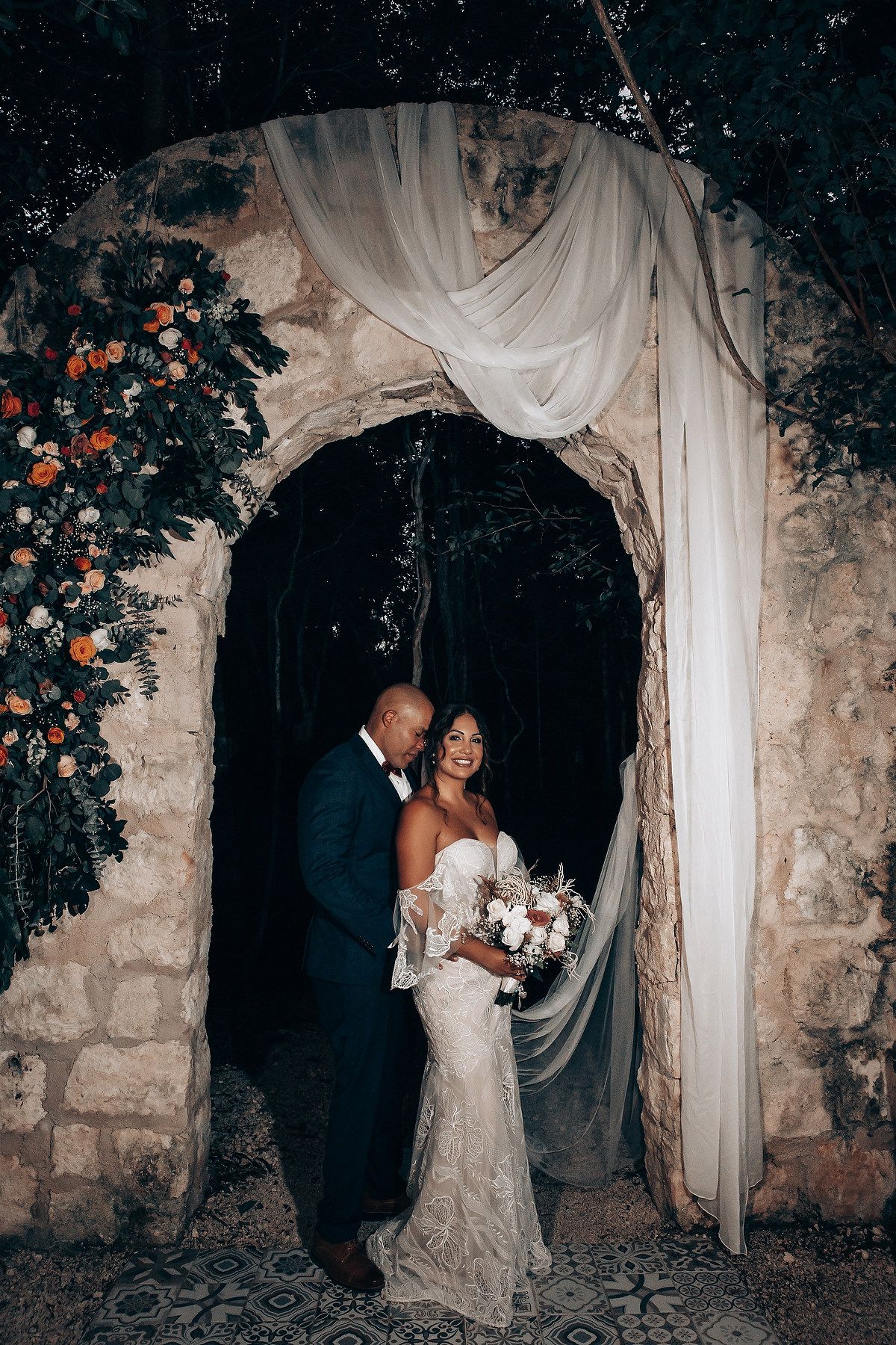 Wedding couple posing in stone archway decorated with white fabric and orange flowers.